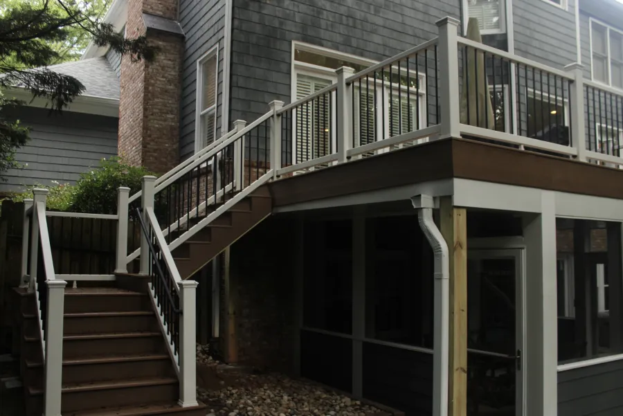 Modern outdoor staircase and balcony with white railing attached to a gray wooden house with stone chimney.