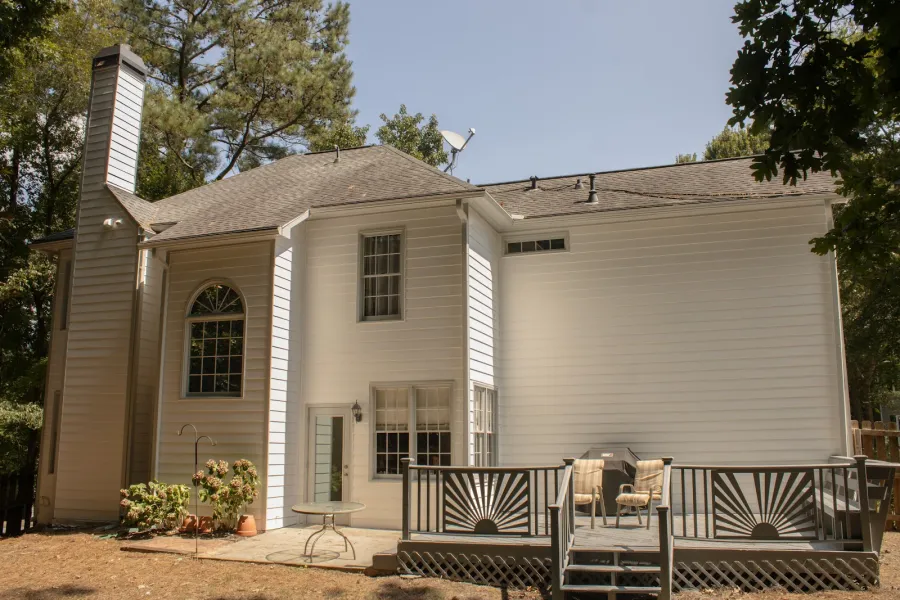 Backyard view of a two-story white house with a deck, outdoor seating, and trees surrounding.