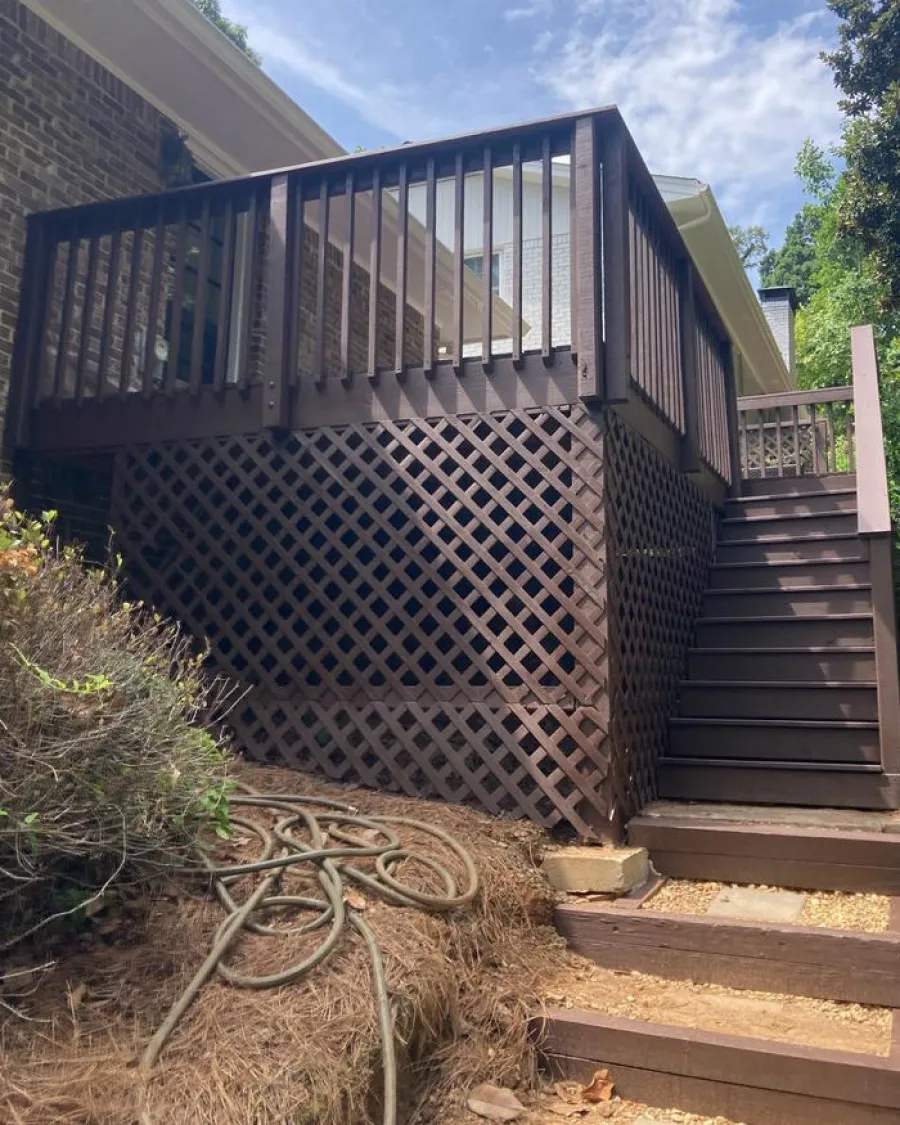 Raised backyard wooden deck with lattice skirt and stairs adjacent to a brick house under blue sky.