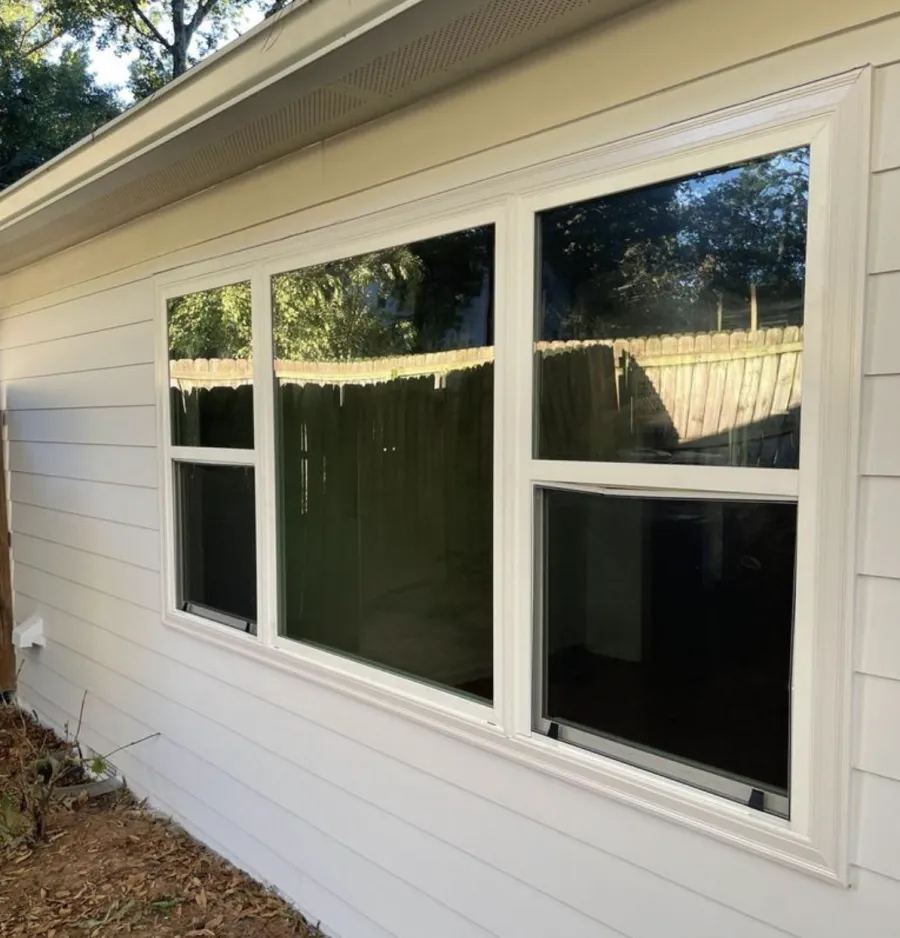 White three-panel house window with sliding sections reflecting backyard wooden fence and trees.