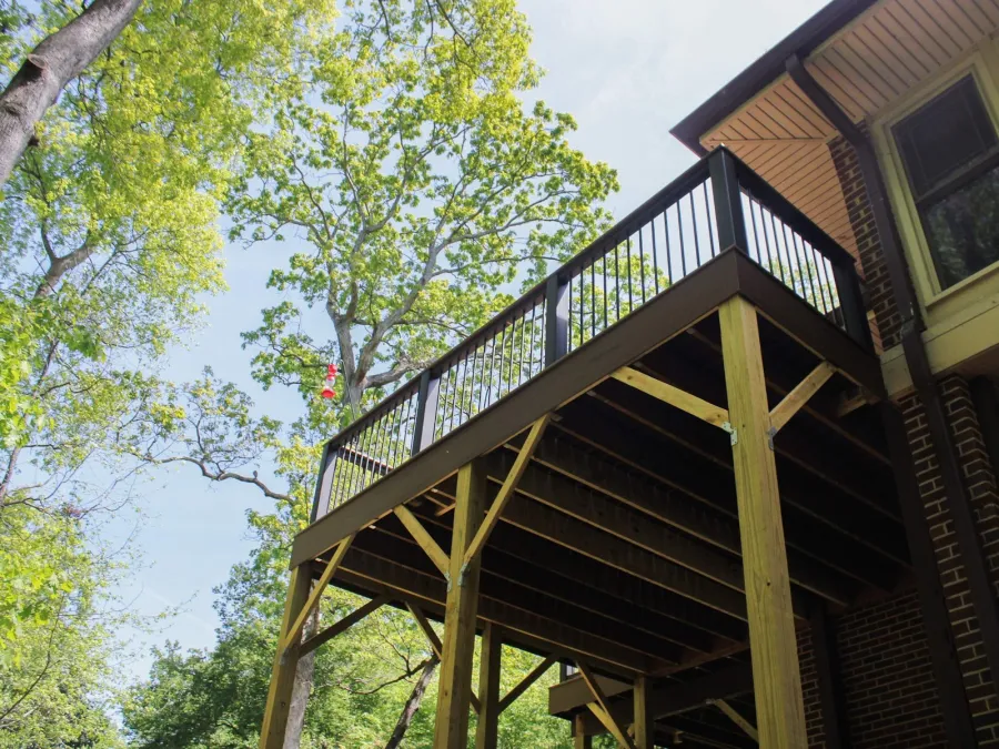 Wooden backyard deck with black metal railing attached to brick house surrounded by green trees under blue sky