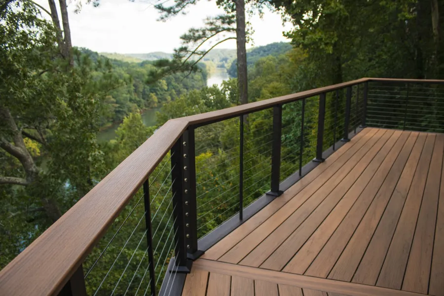 Modern wooden deck with metal cable railing overlooking a lush forest and lake in the distance under a partly cloudy sky.