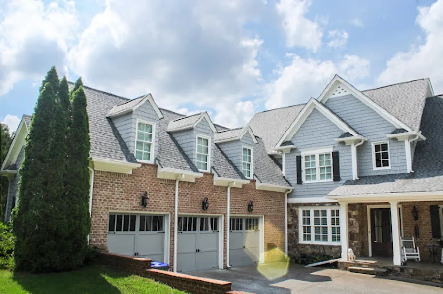 Large suburban house with a brick and blue siding exterior, three-car garage, and tall evergreen trees under a partly cloudy sky