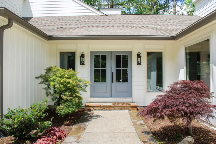 Front entrance of a white house with gray double doors, stone steps, and vibrant green and red foliage.