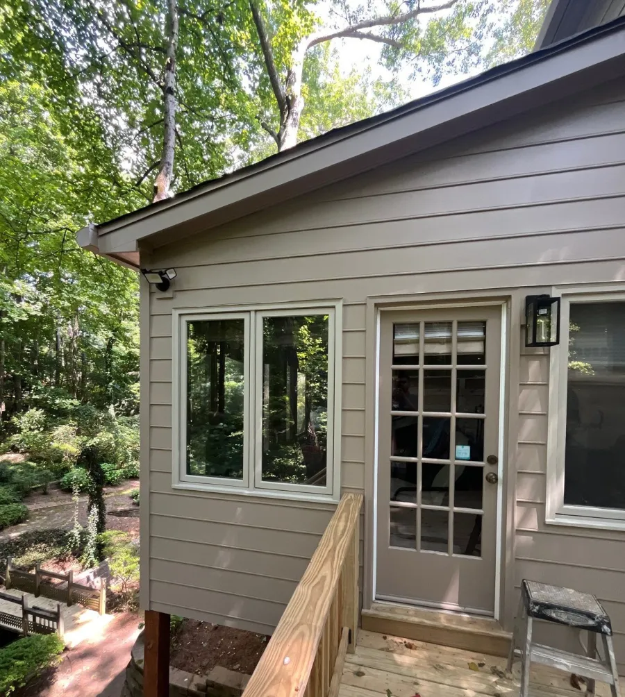 Exterior view of a beige house with glass-paneled door, white-framed windows, and wooden deck surrounded by trees.