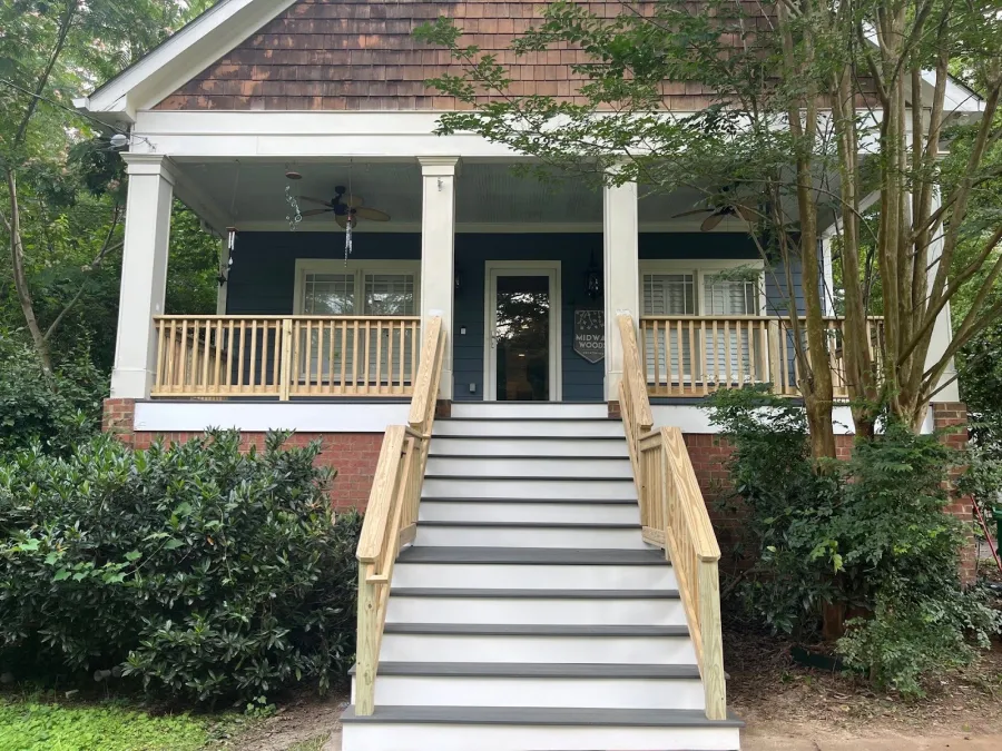 Front view of a porch with wide stairs, wooden railing, brick foundation, and surrounding green shrubs and trees
