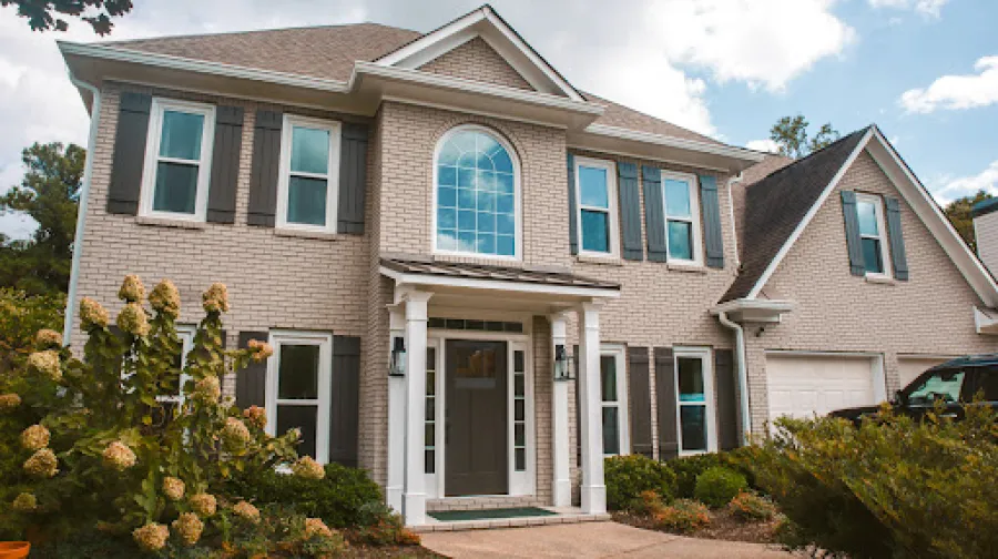 Two-story white house with green roof, front porch, stairs, surrounded by bushes and a clear blue sky.