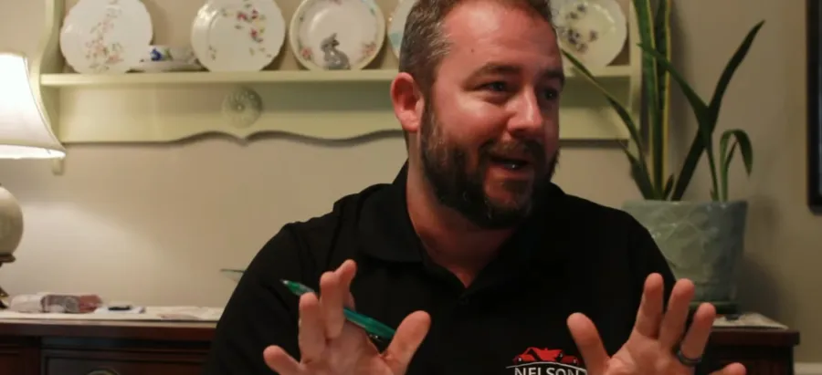 Man with beard gesturing while holding pen in cozy home setting with plates and plant in the background