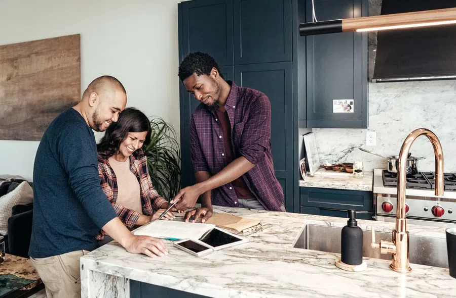 Three people smiling and discussing plans at a modern kitchen island with tablet and documents.