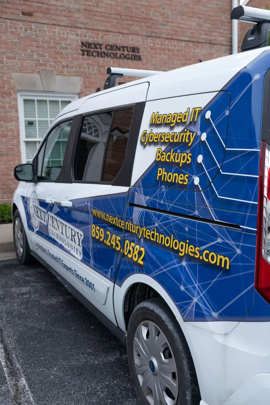 Next Century Technologies service van with IT services and contact info wrapped in blue tech design parked by brick building.