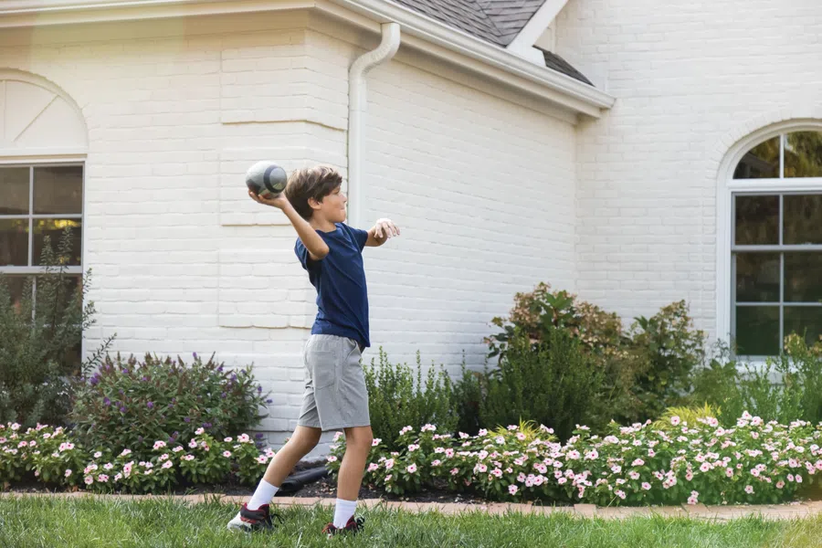 Young boy in navy shirt and gray shorts playing catch with football outside a white brick house.