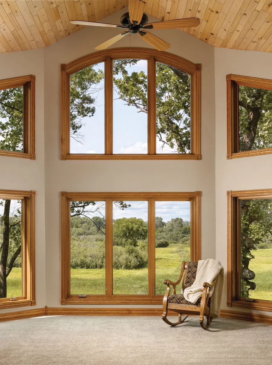Bright room with large wooden windows, a ceiling fan, and a rocking chair overlooking a green landscape.