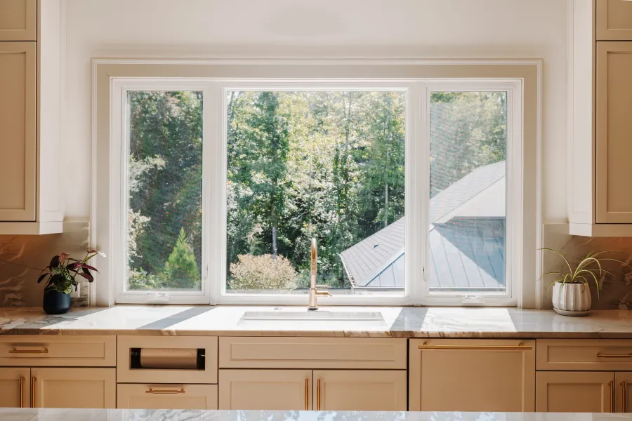 Bright kitchen sink area with large window overlooking green trees and rooftop outside.
