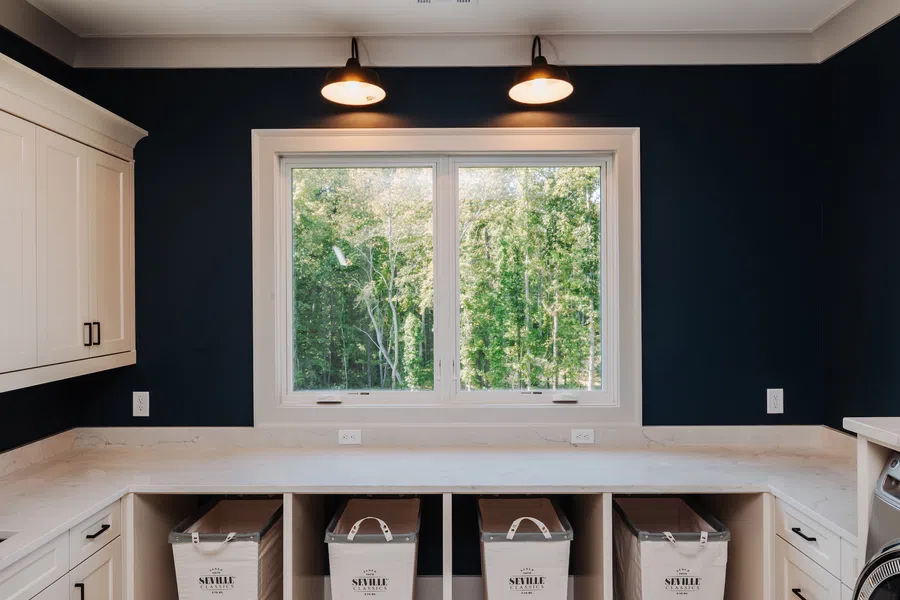Bright laundry room with white cabinets, marble countertop, three labeled storage bins, and window showing green trees.