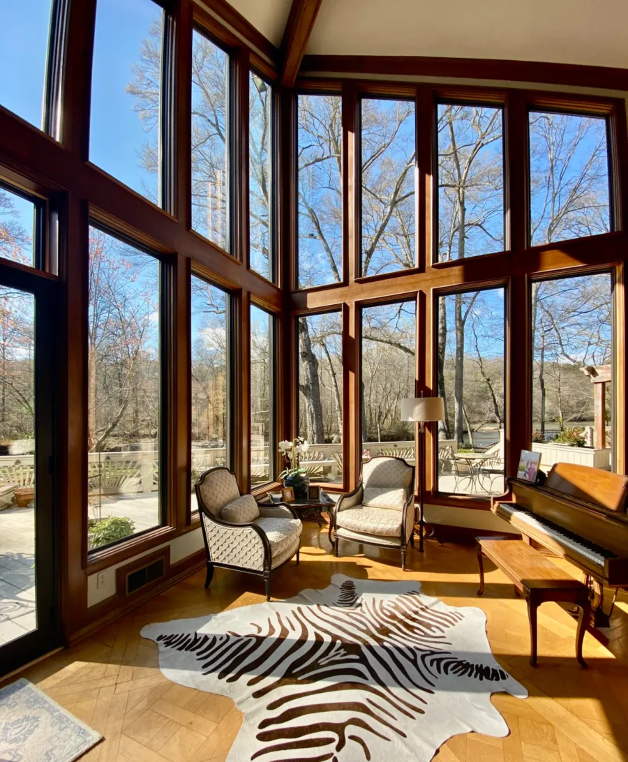 Sunlit room with large floor-to-ceiling windows, two armchairs, zebra-patterned rug, and wooden piano.