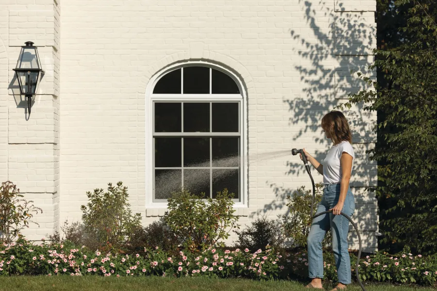 Woman watering garden flowers with hose in front of white brick house with arched window.