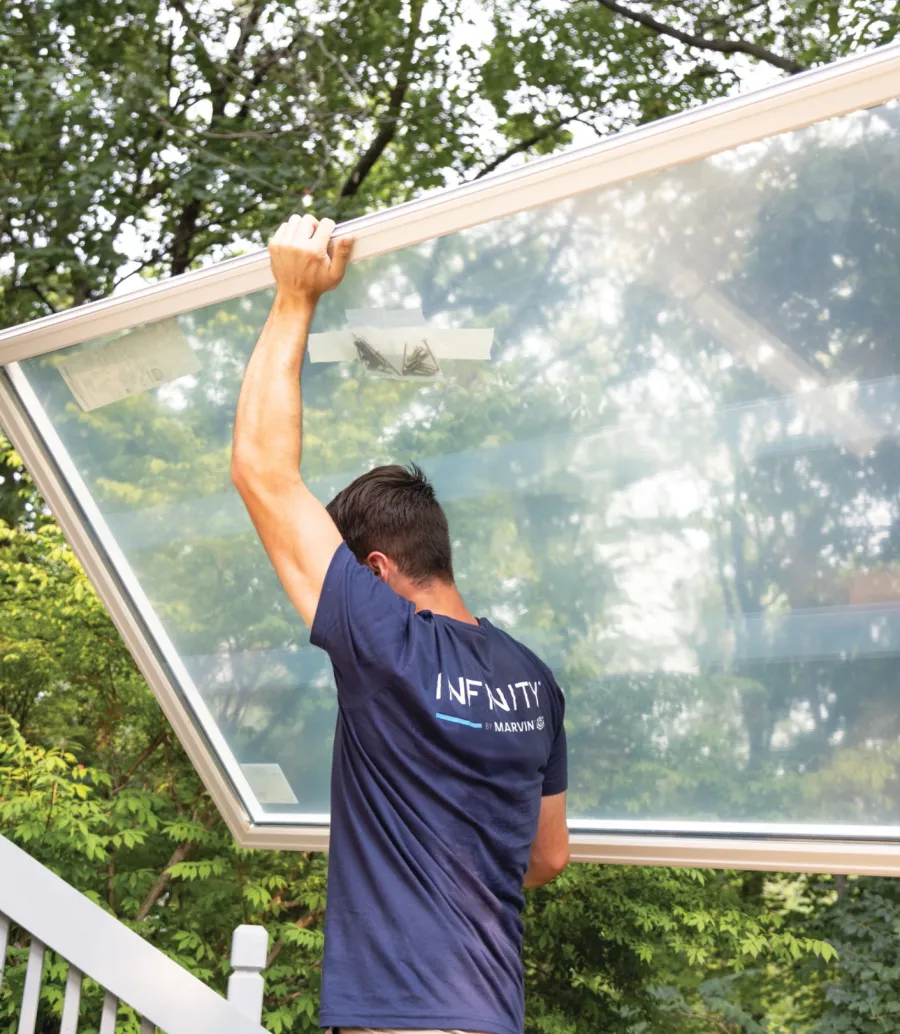 Man in infinity by Marvin shirt installing a large window panel outdoors surrounded by green trees.