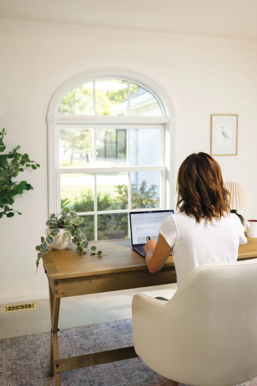 Woman working on laptop at wooden desk in a bright room with large arched window and indoor plants.