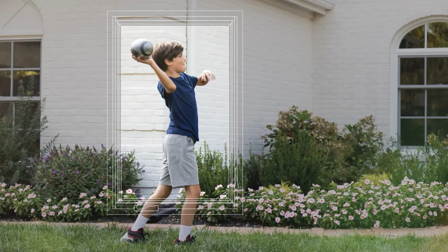 Young boy in blue shirt and gray shorts throwing a football in a garden with flowers and white brick house.