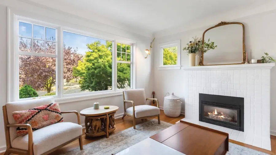 Bright living room with large windows, two armchairs, wooden tables, fireplace, and decorative plants.