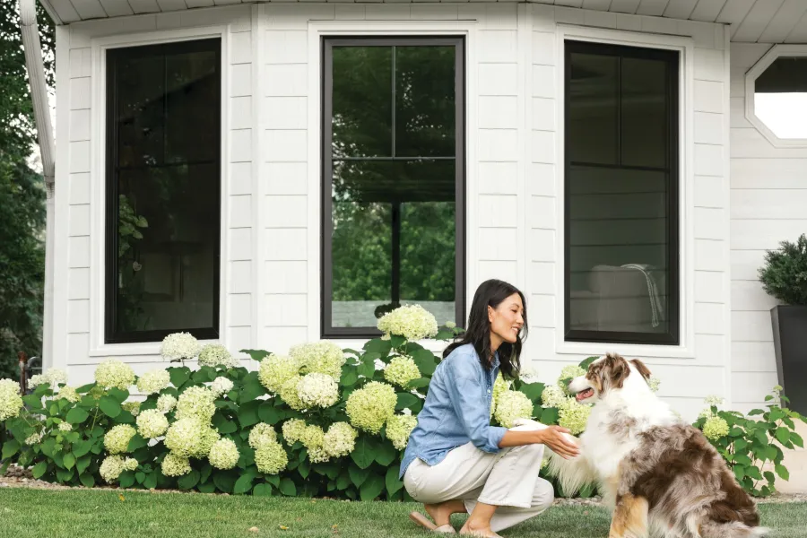 Woman watering garden flowers with hose in front of white brick house with arched window.