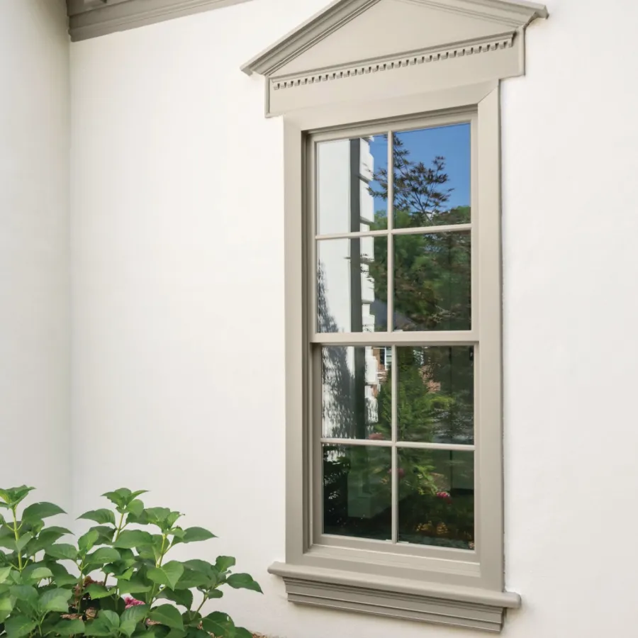 Traditional beige-framed window with ornate pediment and lush green shrub with pink flowers below