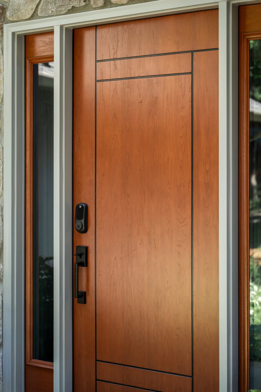 Modern wooden front door with black handle and smart lock beside tall narrow windows in stone exterior