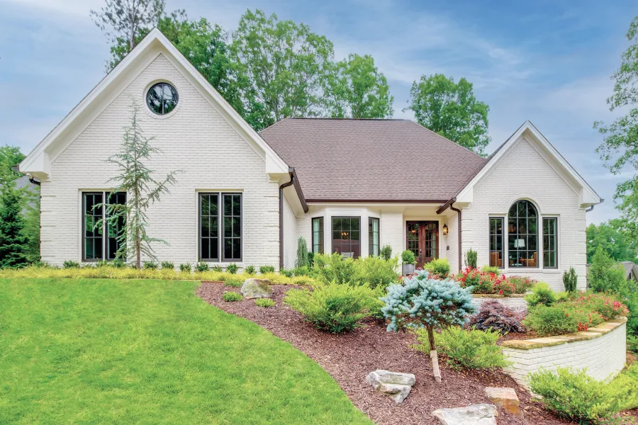 White brick house with steep gable roof, large windows, and landscaped garden with shrubs and flowers.