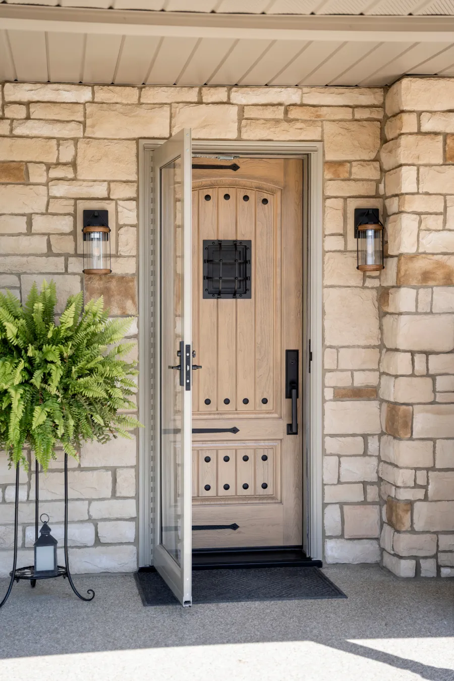 Front wooden door with decorative black hardware and glass storm door in stone house entryway with lanterns and fern plant.