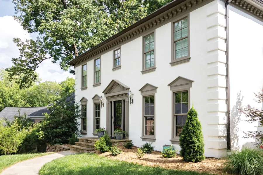 a white house with trees and bushes and tan trim around the roofline, front entry, and upper and lower level double hung Infinity from Marvin fiberglass windows.