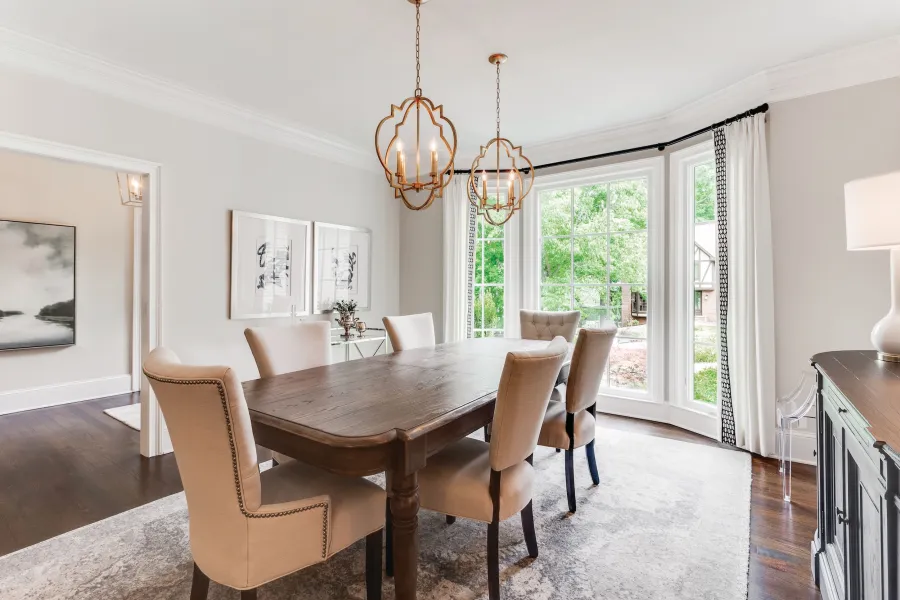 Bright modern dining room with wooden table, beige upholstered chairs, decorative lighting, and large bay window.