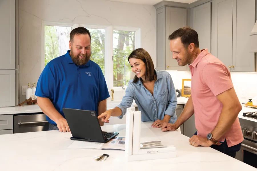 Three people smiling and discussing something on a laptop in a modern kitchen with white countertops