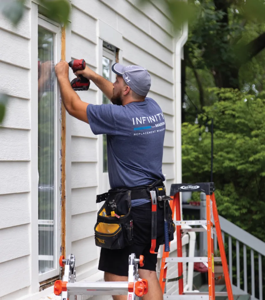 Worker installing replacement window on house exterior using power drill with ladder and tool belt