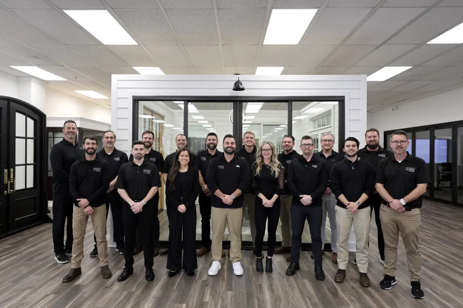 Professional team of 16 men and women posing indoors in office environment, wearing black shirts and khakis.