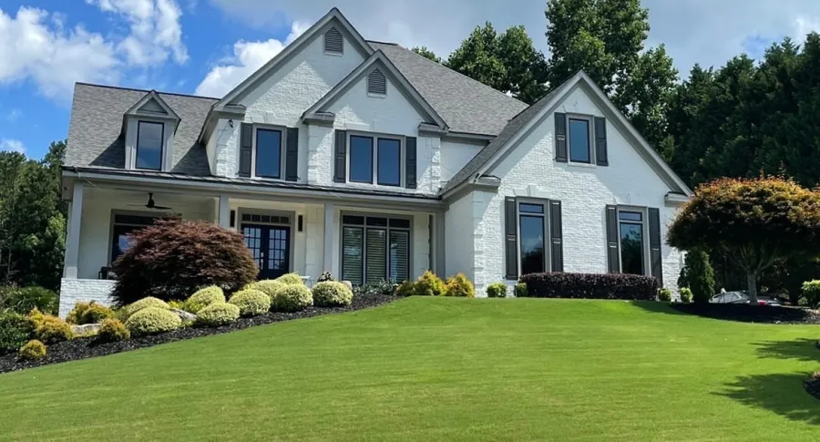 Large white brick house with grey roof, multiple windows, and manicured lawn under blue sky with clouds.