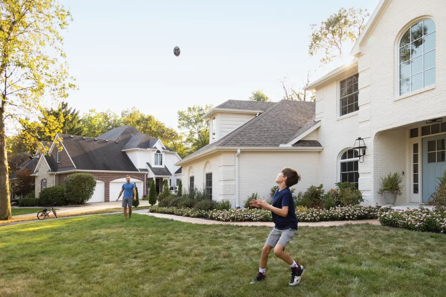 Boy catching football in front yard while adult watches near a suburban house on a sunny day