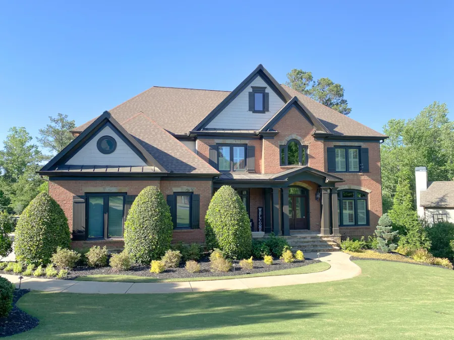 Two-story brick house with a large front lawn, shrubs, and a clear blue sky on a sunny day.