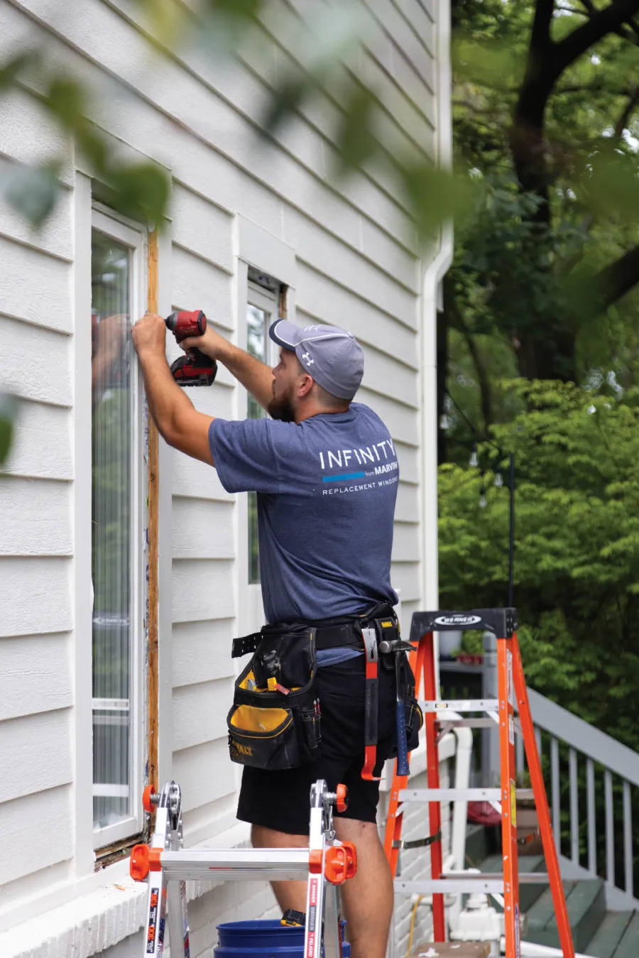 Worker installing replacement window on house exterior using power drill with ladder and tool belt