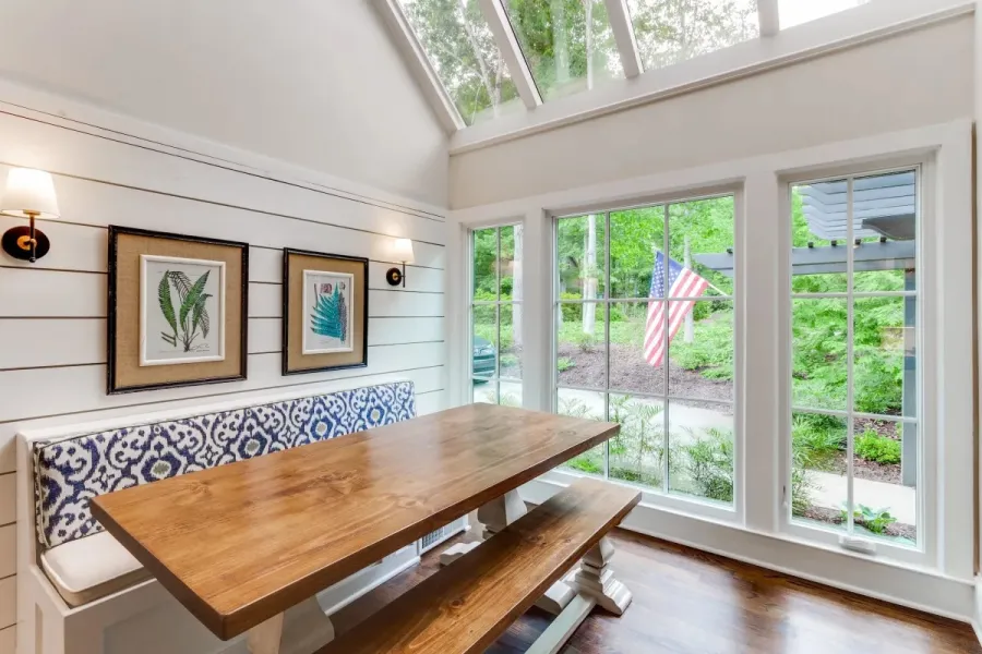 Bright dining nook with wooden table, patterned bench, botanical art, and large windows showing greenery and flag.