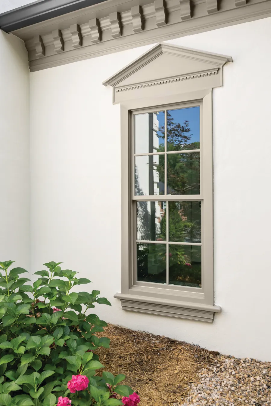 Classic beige window with pediment trim on white wall next to garden with green plants and pink flowers.
