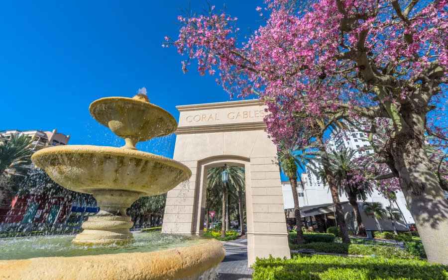 Coral Gables arch entrance with flowing stone fountain and pink flowering tree under clear blue sky.