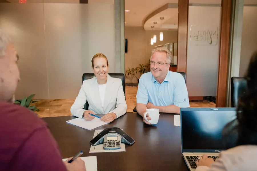 Two professionals in a meeting room smiling, seated at a table with a conference phone and a laptop