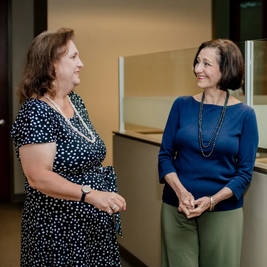 Two professional women smiling and chatting in a modern office corridor with glass partitions.
