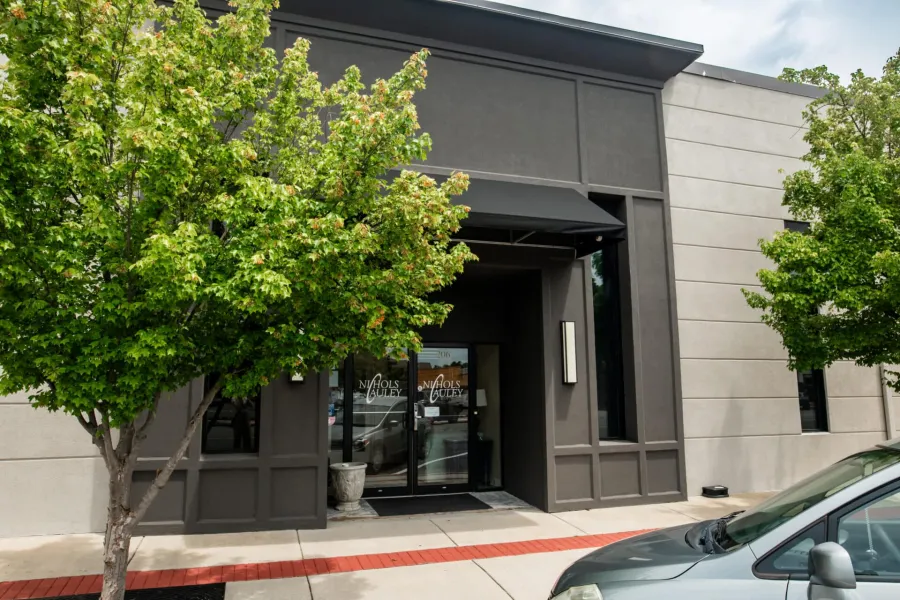 Modern commercial building entrance with glass doors, awning, and green trees on sidewalk under blue sky