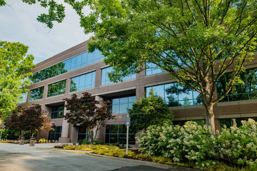 Modern office building with large glass windows surrounded by lush green trees and landscaping on a sunny day