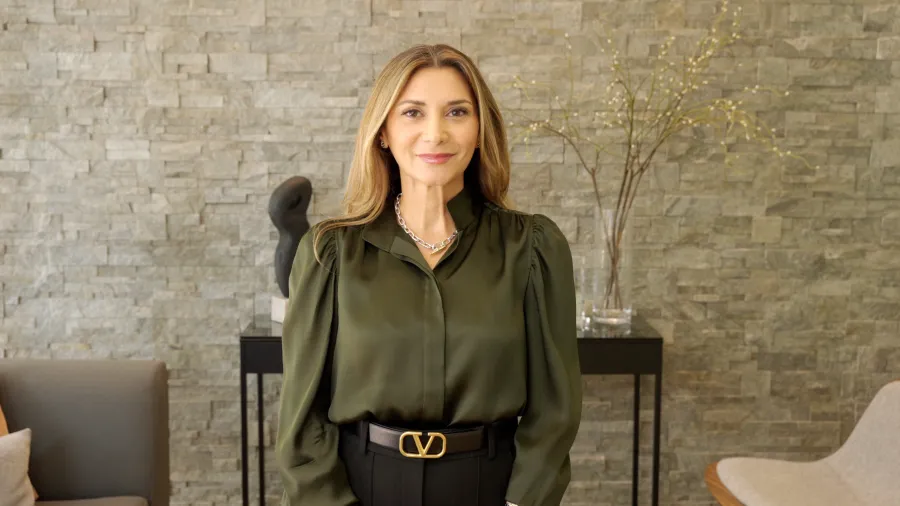 Smiling woman wearing olive green blouse and black pants standing in modern living room with stone wall background.