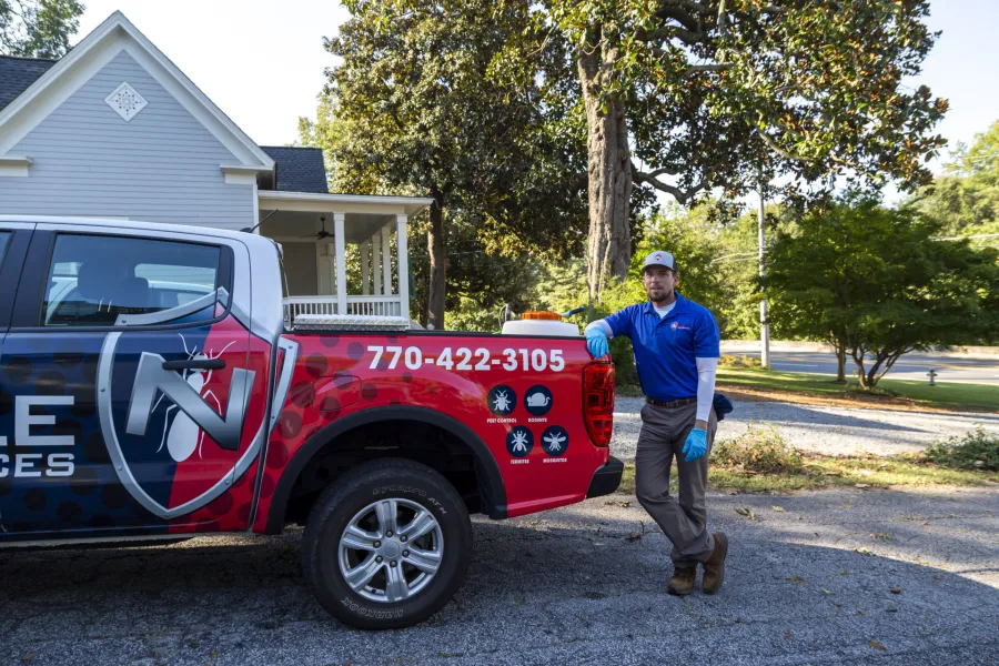 Pest control professional in uniform leaning on branded service truck in residential neighborhood on sunny day