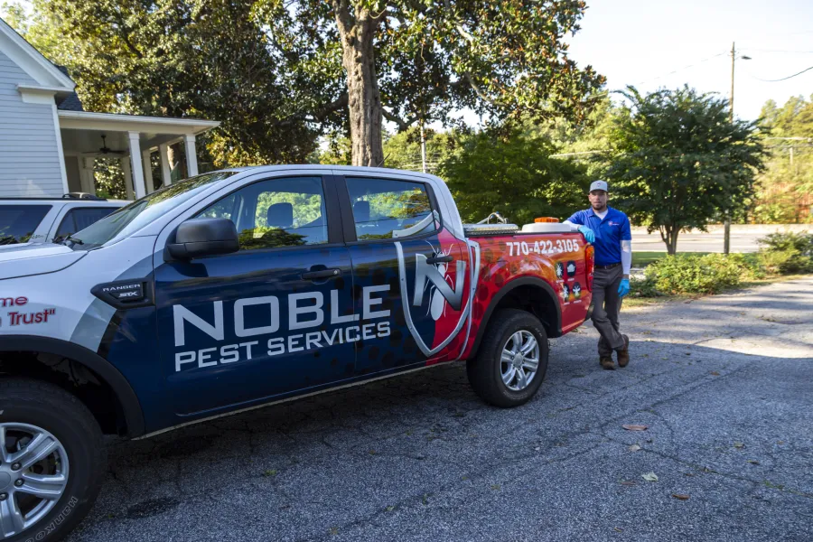 Noble Pest Services branded truck parked outside with a uniformed technician standing beside it on a sunny day.
