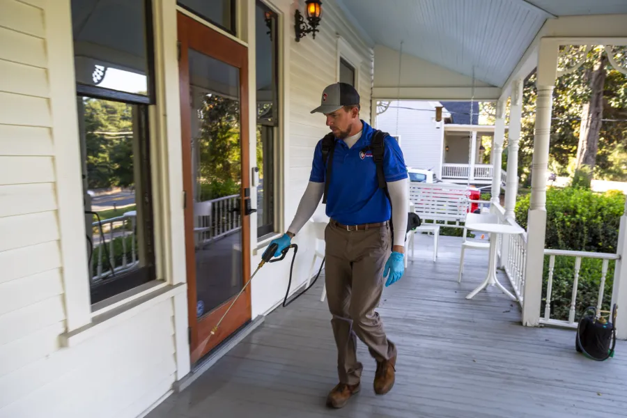 Pest control technician sprays pesticide on a porch of a residential home wearing gloves and uniform.