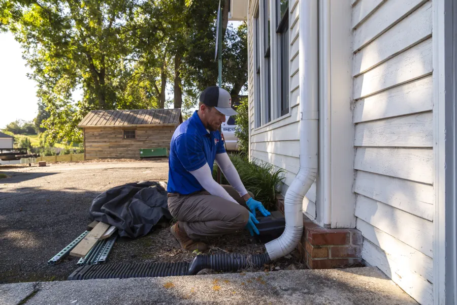 Worker in blue shirt repairing a white house downspout drainage system on a sunny day outdoors.